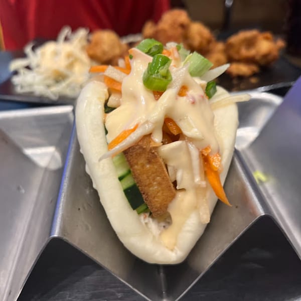 A Senzu Noodle Bar bao bun with fried chicken, vegetables, and sauce in a metal holder.