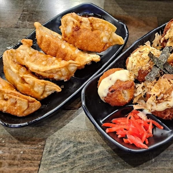 Two black plates of Japanese appetizers, one with fried gyoza and the other with takoyaki and pickled ginger.
