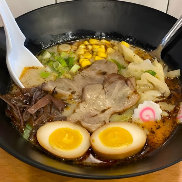 Close-up of a bowl of Senzu Noodle Bar ramen with pork, corn, scallions, fish cake, and two soft-boiled eggs.
